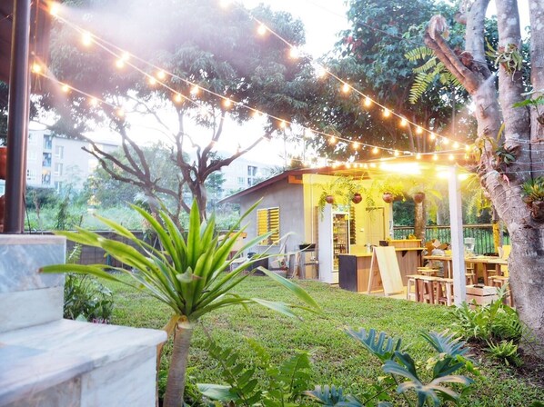 An outdoor seating area with string lights, wooden tables, and chairs, surrounded by greenery and trees.