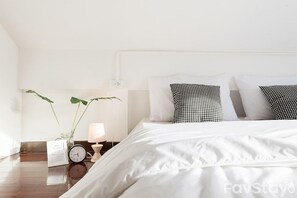 A neatly made bed with white bedding, a checkered pillow, a bedside table with a lamp, and a clock.