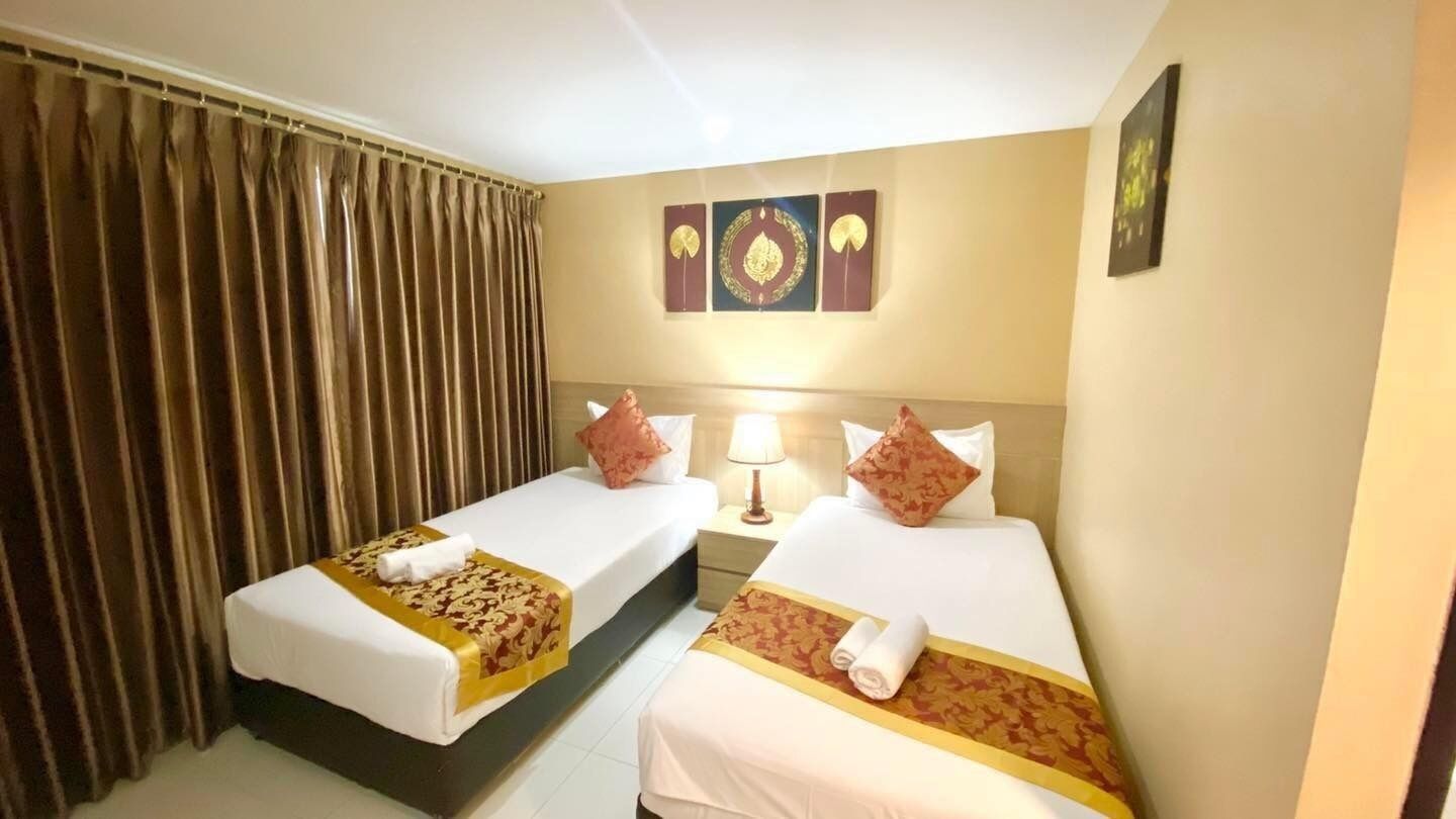 Habitación de hotel con dos camas, cada una con una colcha blanca y un ribete dorado decorativo. Hay una cortina en un lado y una lámpara de pared entre las camas.