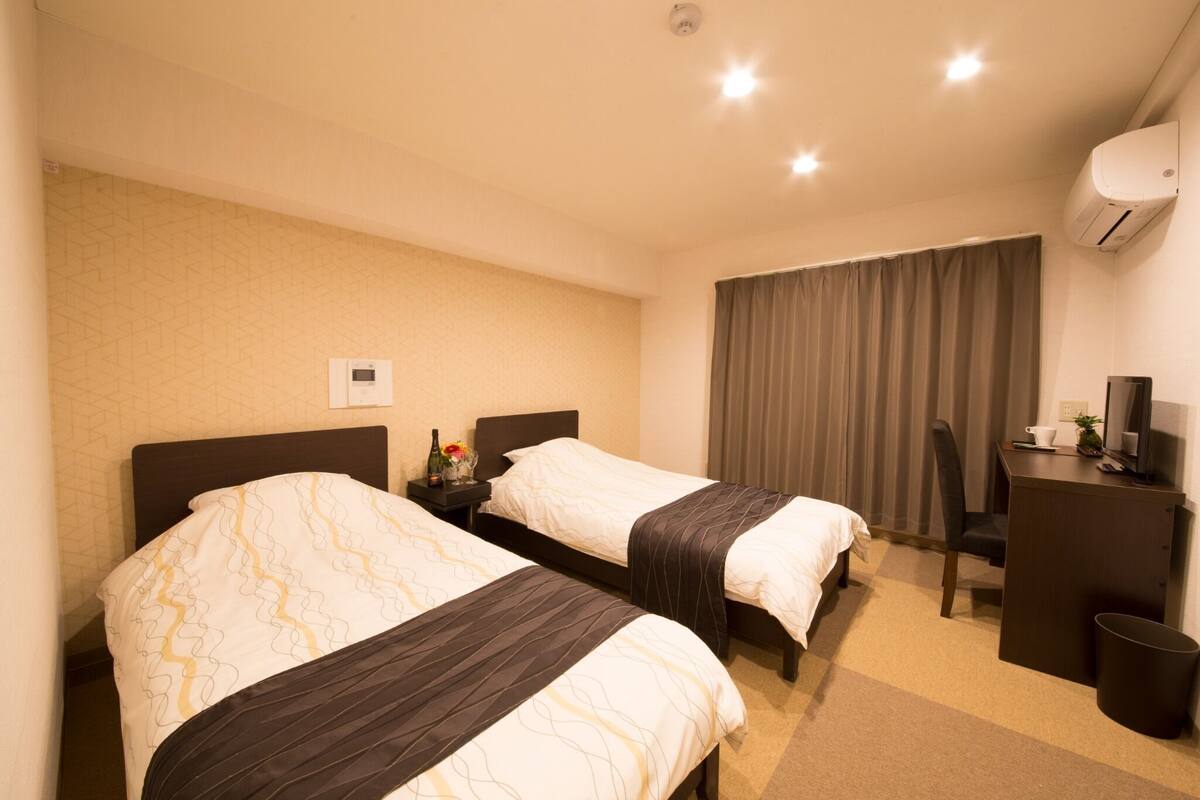 A hotel room with two beds, a desk, a chair, a trash bin, and an air conditioning unit.