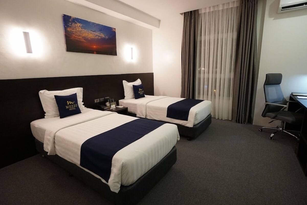 A hotel room with two beds, a desk, a chair, and a painting on the wall.
