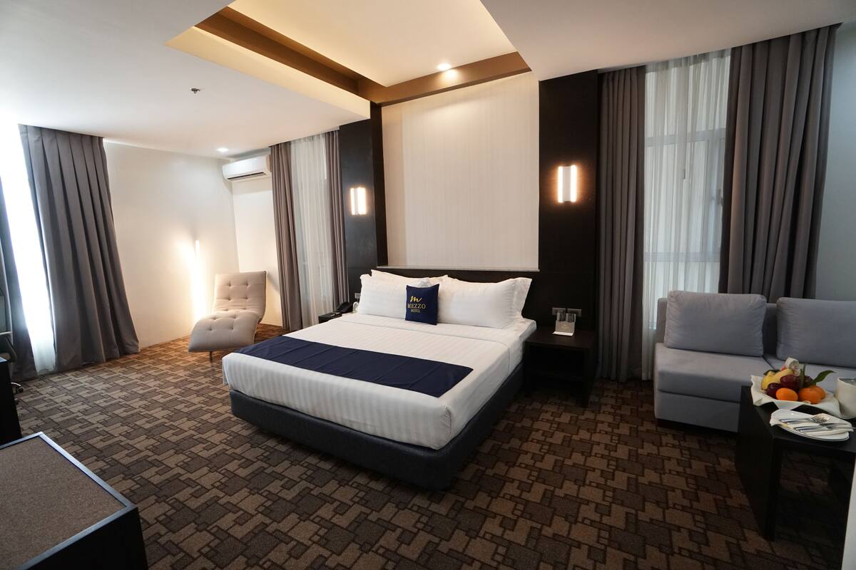Quarto de hotel com uma cama grande, um sofá, uma escrivaninha com uma tigela de frutas e uma cadeira.