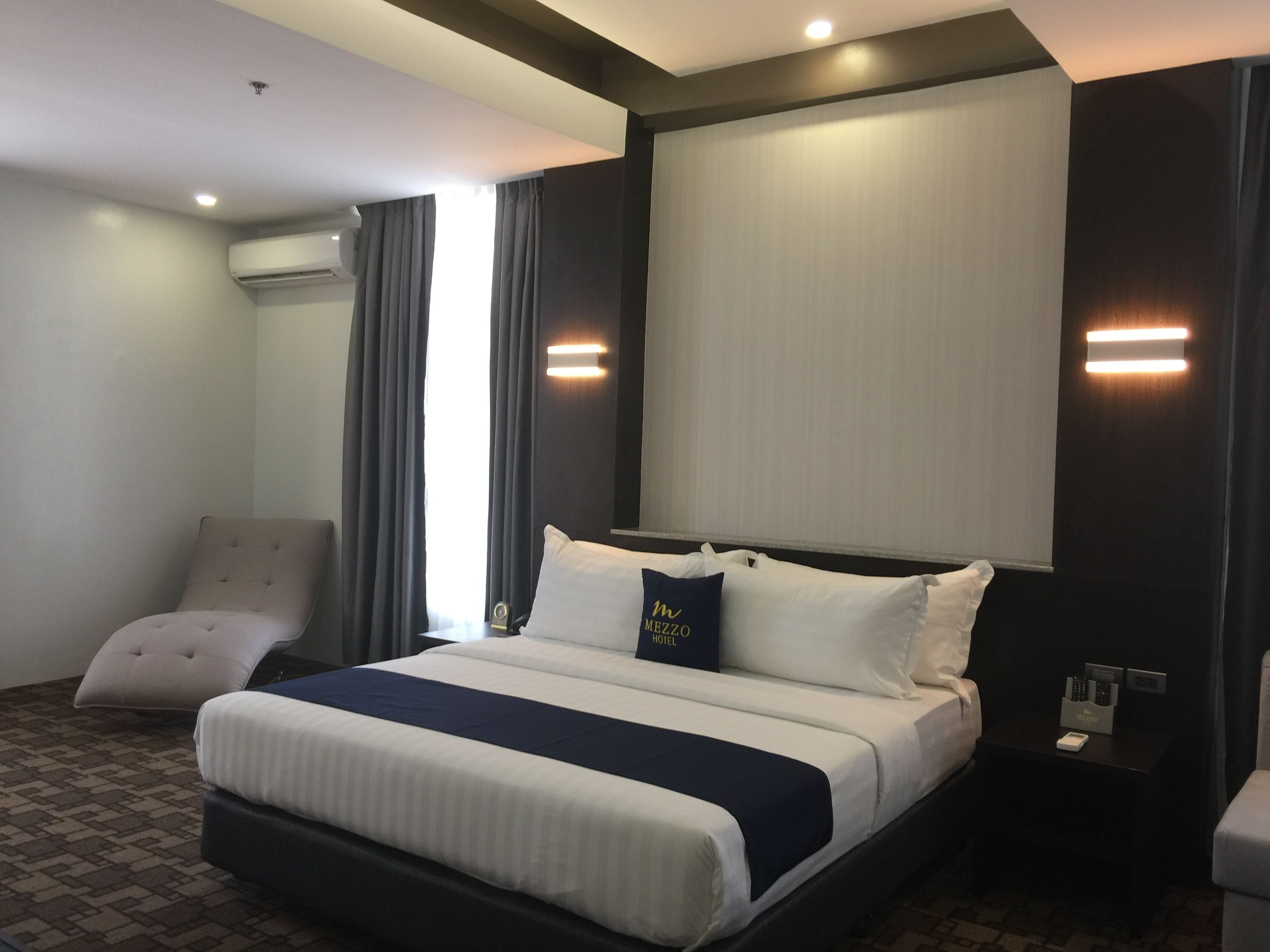 A hotel room with a large bed, a chair, a nightstand, and a window with curtains.