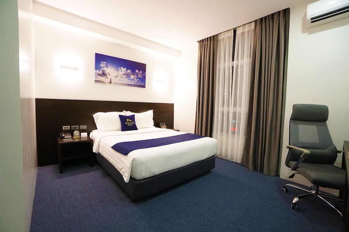 Quarto de hotel com uma cama grande, uma escrivaninha e uma cadeira. Há um quadro na parede e um aparelho de ar condicionado acima da cama.