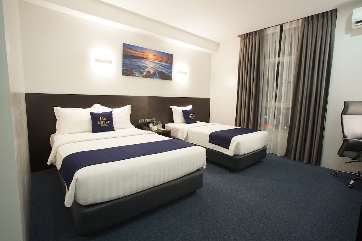 Quarto de hotel com duas camas, uma mesinha de cabeceira, uma cadeira, um quadro com uma paisagem de pôr do sol e uma janela grande com cortinas.