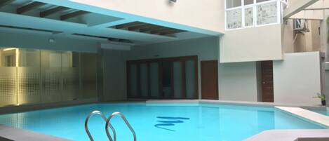 An indoor swimming pool with clear blue water, a diving board, and a modern architectural design with a high ceiling and large windows.