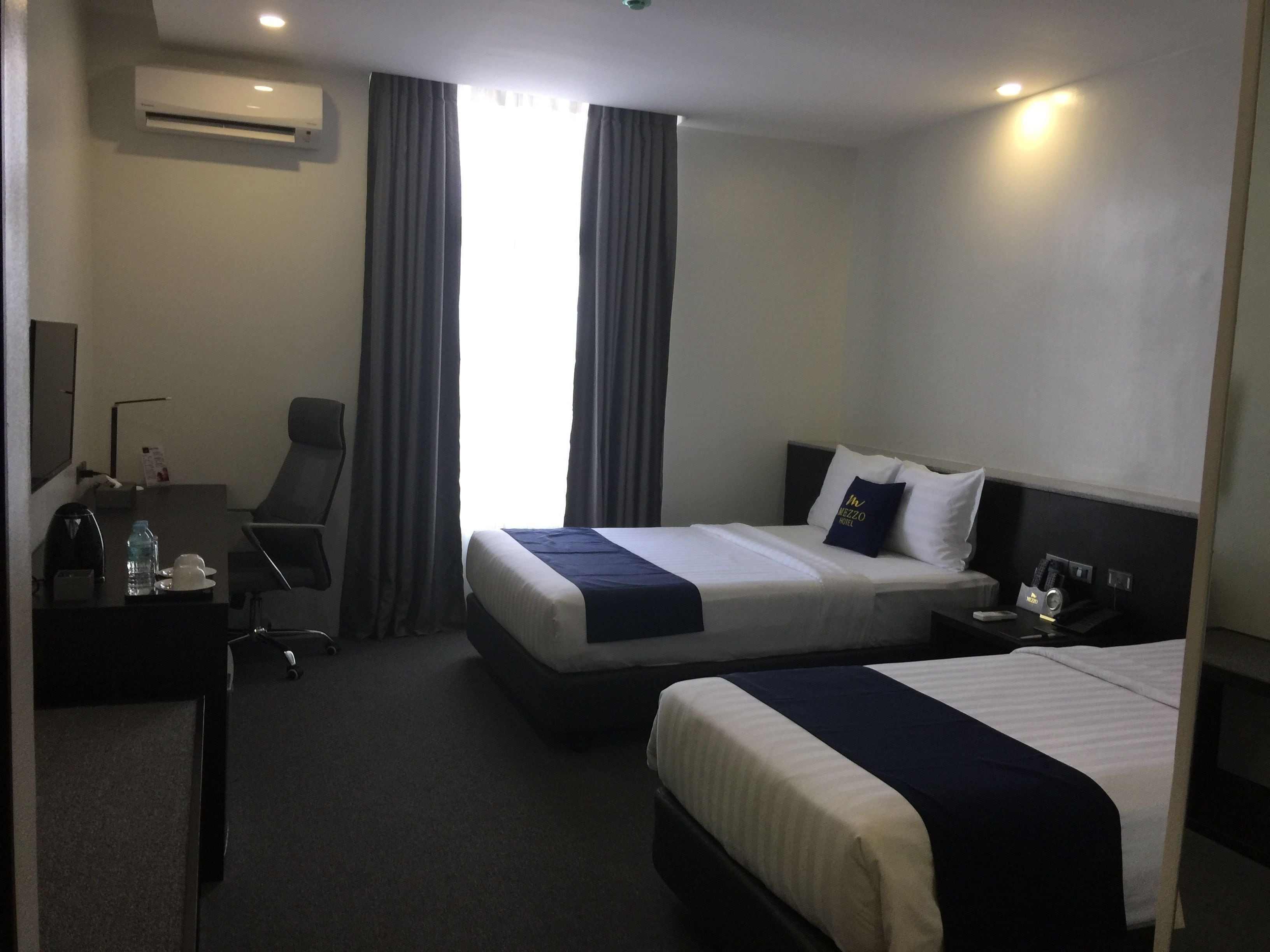 Habitación de hotel con dos camas, un escritorio con una silla, una lámpara de escritorio, una botella de agua, una taza y un minibar.