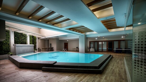 Une piscine intérieure moderne dotée d’une terrasse en bois, d’un mur en verre et d’un plafond avec des poutres en bois.