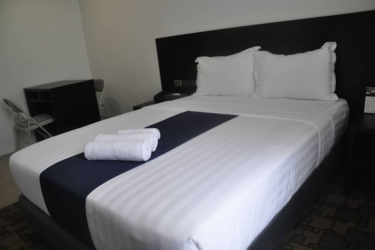 Uma cama bem arrumada com lençóis brancos e detalhes em azul escuro, uma mesa de cabeceira com abajur e uma cadeira em um quarto de hotel.