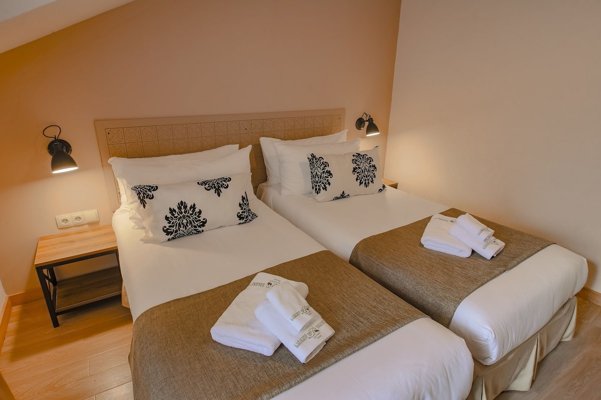 Quarto de hotel com duas camas, cada uma com cabeceira, abajures e toalhas dobradas com cuidado.