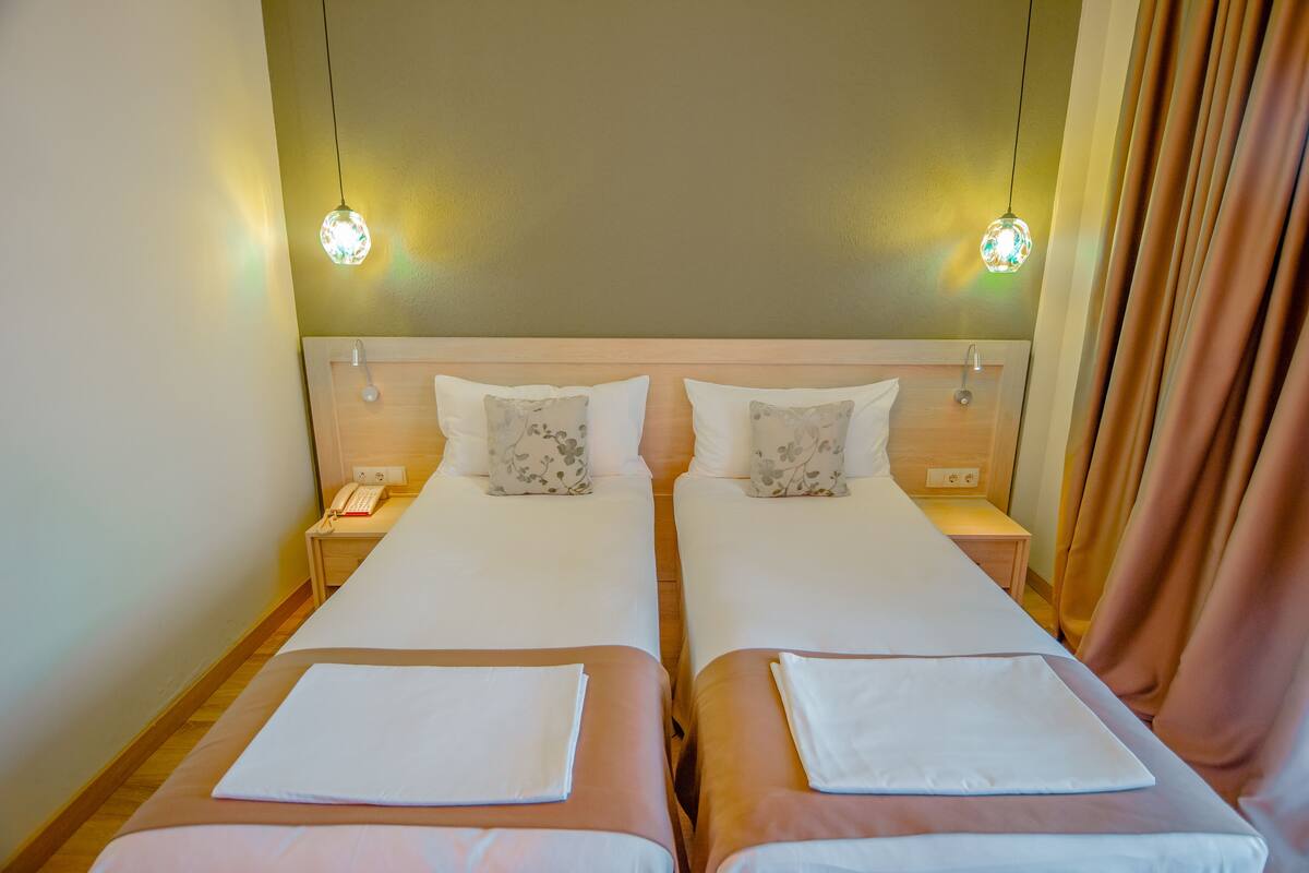 Quarto de hotel com duas camas, cada uma com um cobre-leito branco e uma almofada. Há mesas de cabeceira com abajures e um telefone.