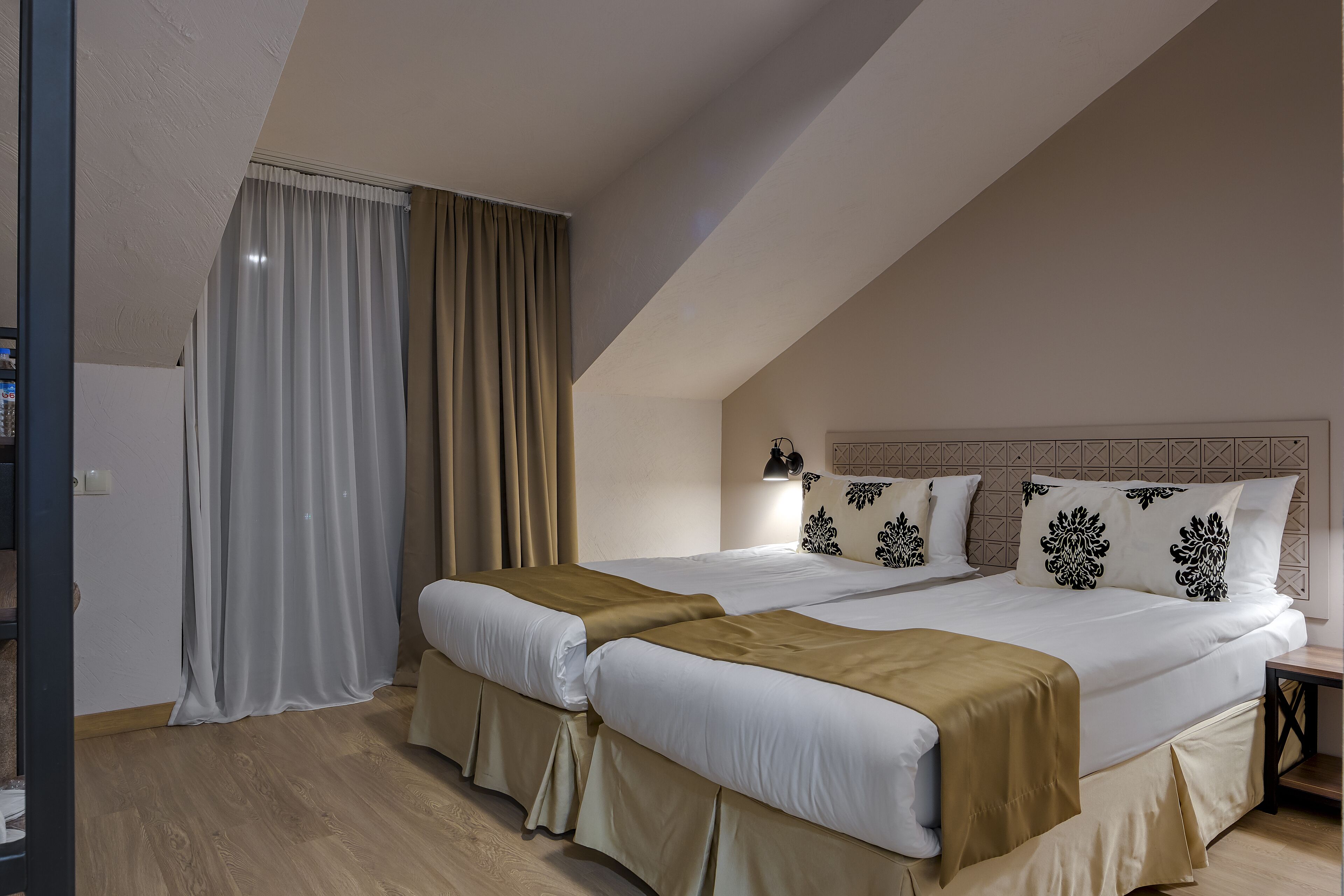 Un dormitorio con dos camas, ropa de cama color beige, piso de madera y una mesita de noche con lámpara.