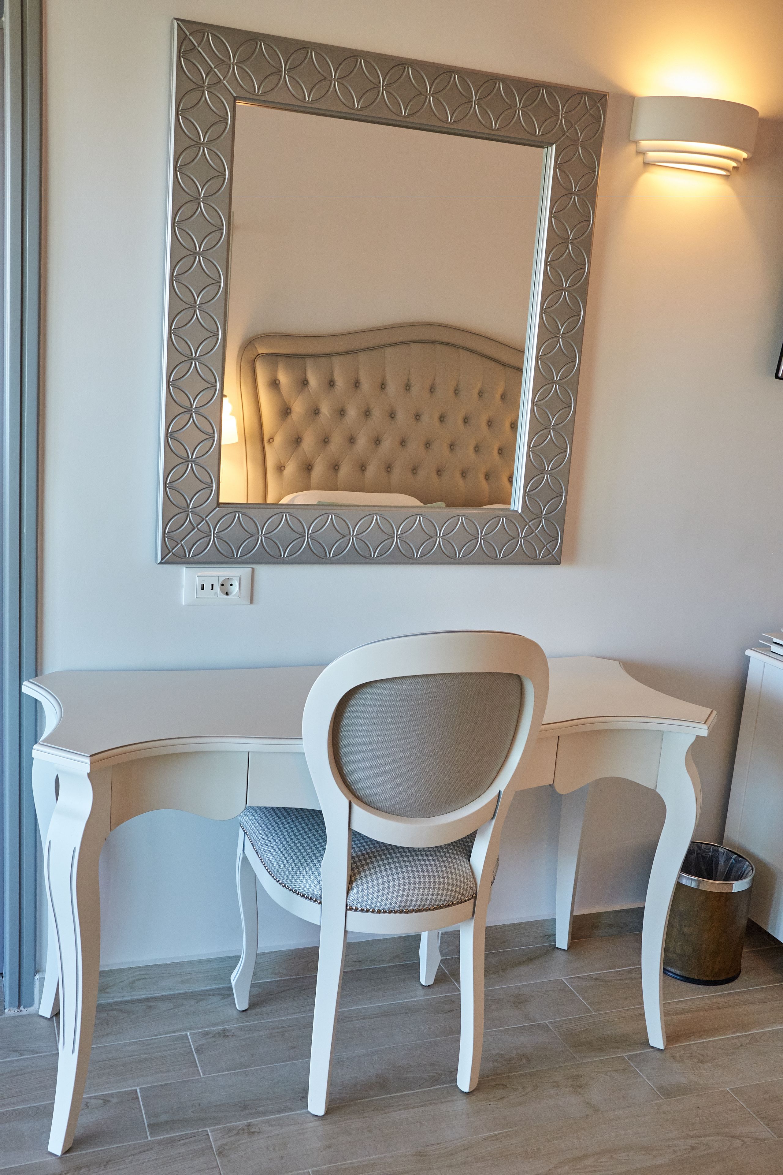 Una camera moderna con una scrivania bianca, una testiera imbottita e uno specchio con cornice geometrica fissato alla parete.