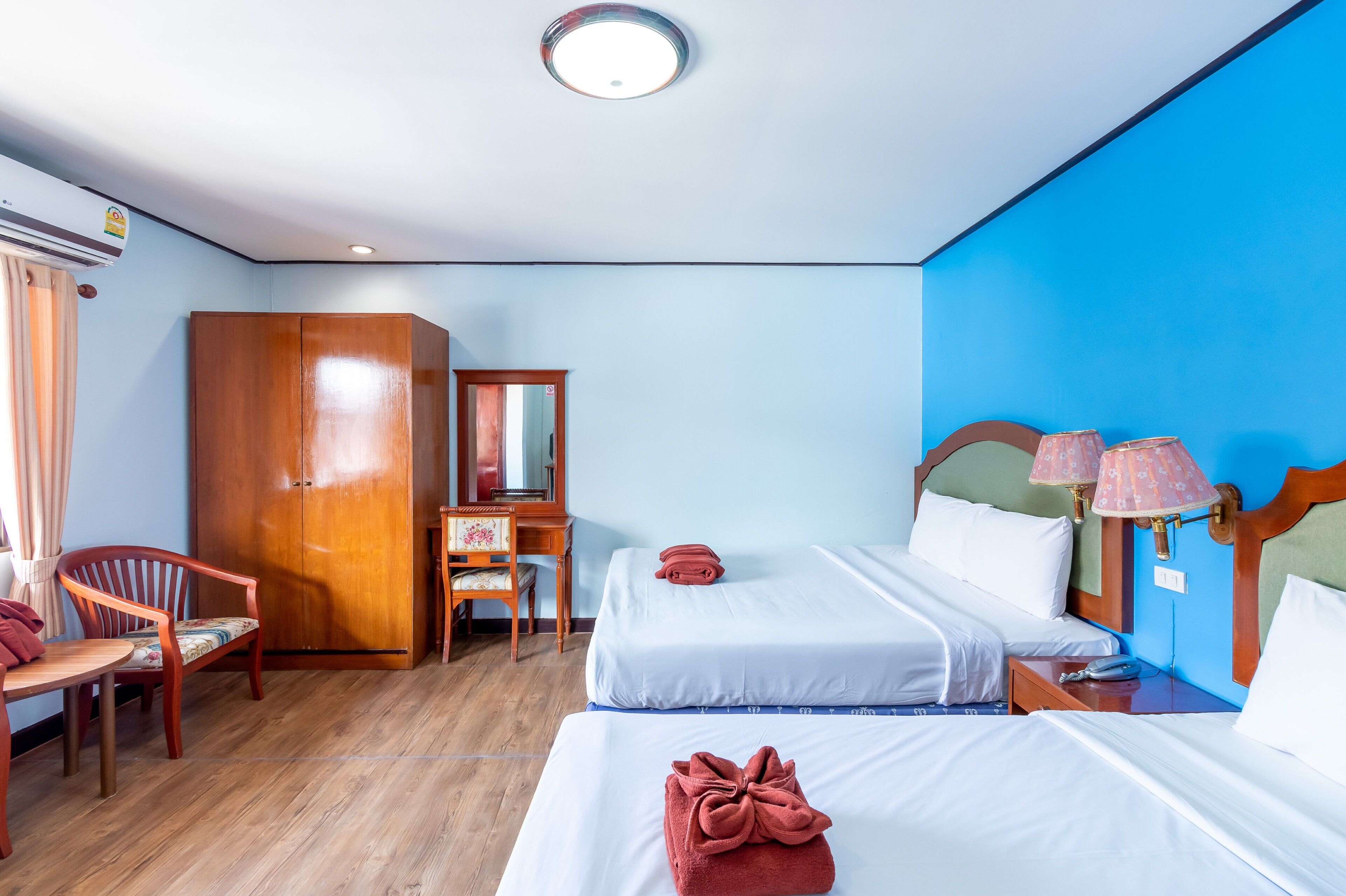 A hotel room with two beds, a wooden wardrobe, a desk with a chair, and a desk lamp.