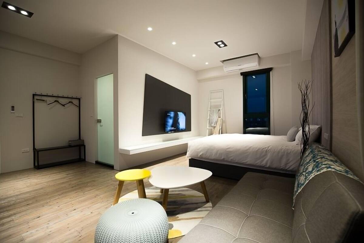 Un dormitorio moderno con cama, televisor montado en la pared, un pequeño rincón de descanso y una ventana con vistas al cielo nocturno.