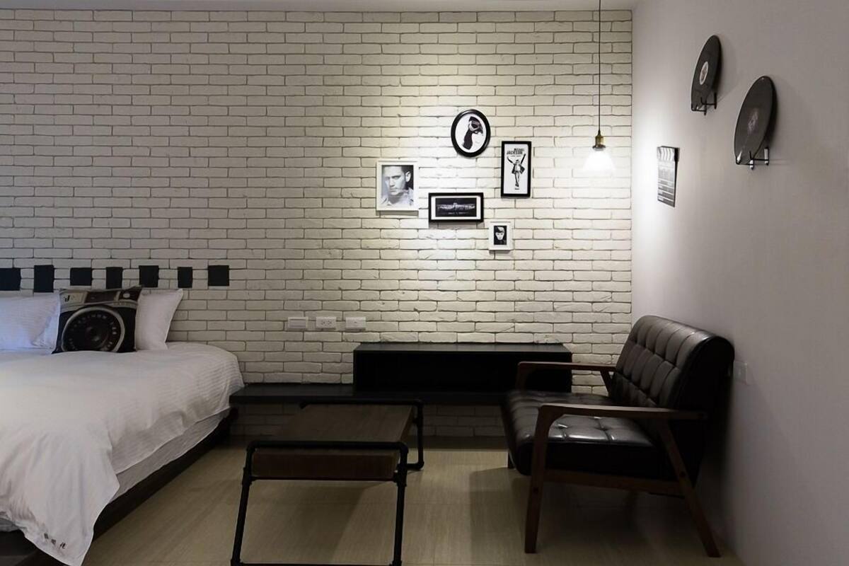 Un dormitorio con pared de ladrillo blanco, una cama, una mesita, un sillón de cuero y una obra de arte colgada en la pared.