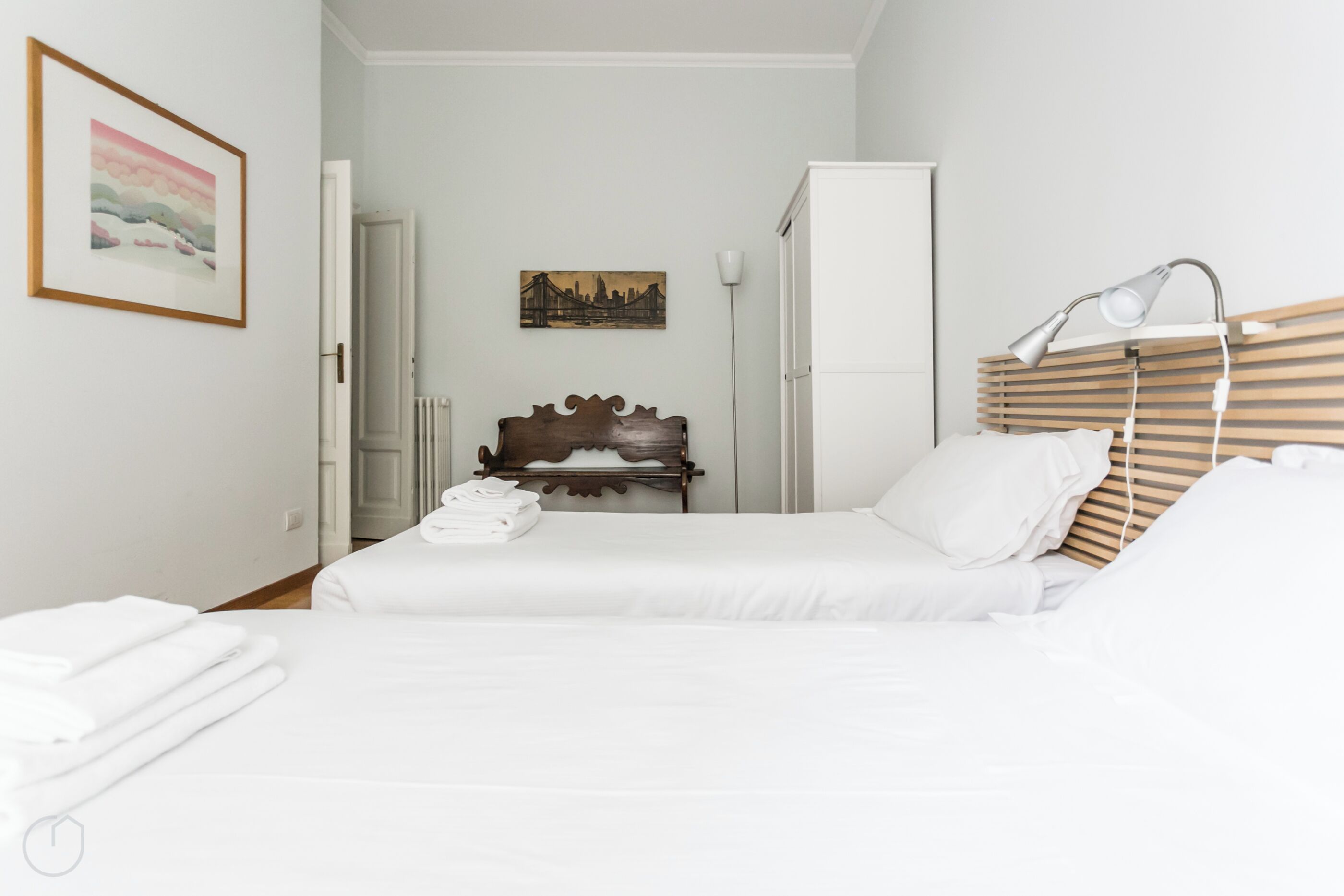 Una habitación de hotel con una cama, cabecera de madera, lámpara de noche, armario blanco y un cuadro en la pared.