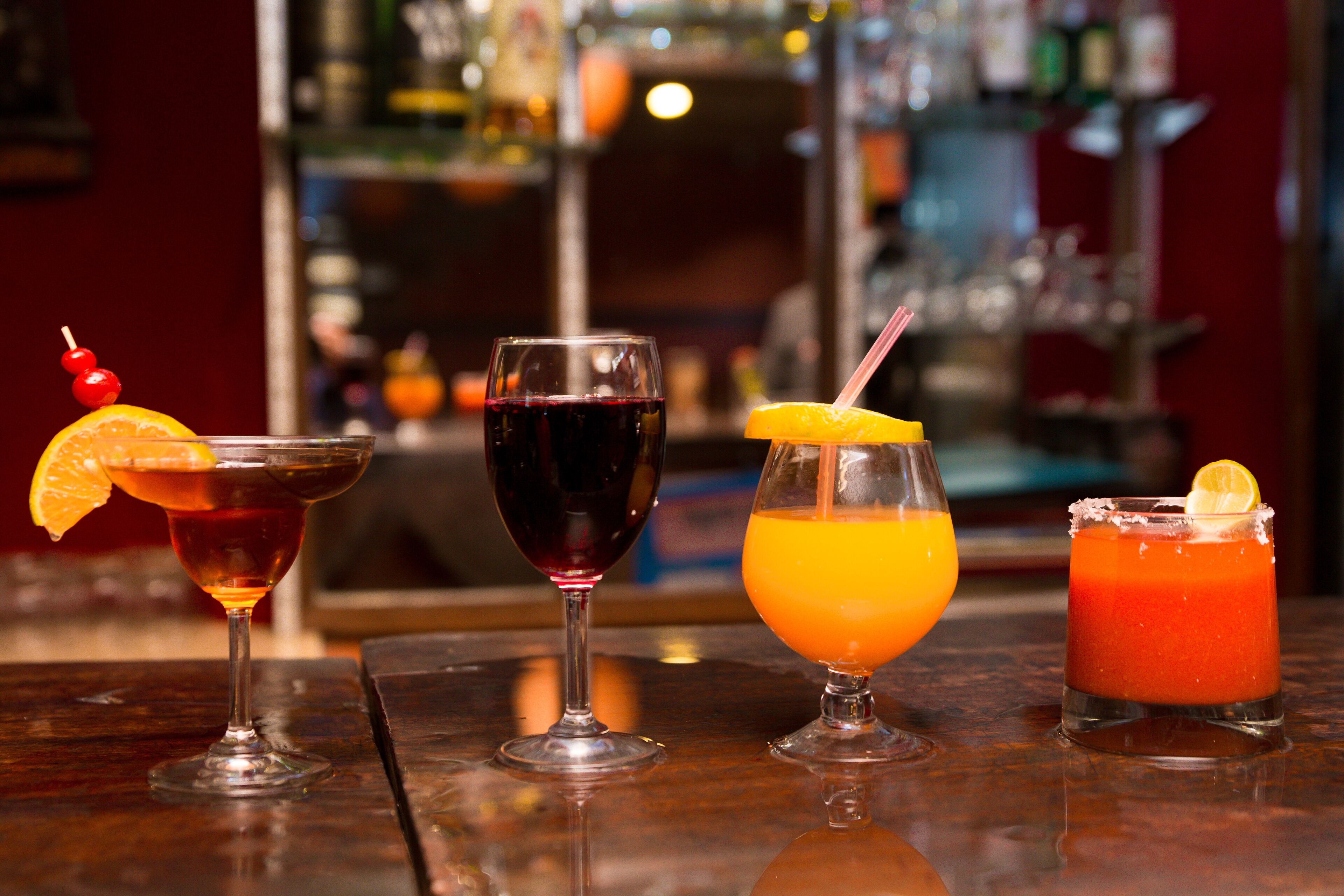 Quatre cocktails différents sont présentés sur le comptoir d’un bar, avec un arrière-plan flou composé d’étagères et de bouteilles.