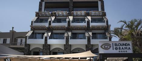 A multi-story hotel with balconies, a sign indicating 'ELOUNDA ORAMA', and a 'JOY!' sign on the facade.