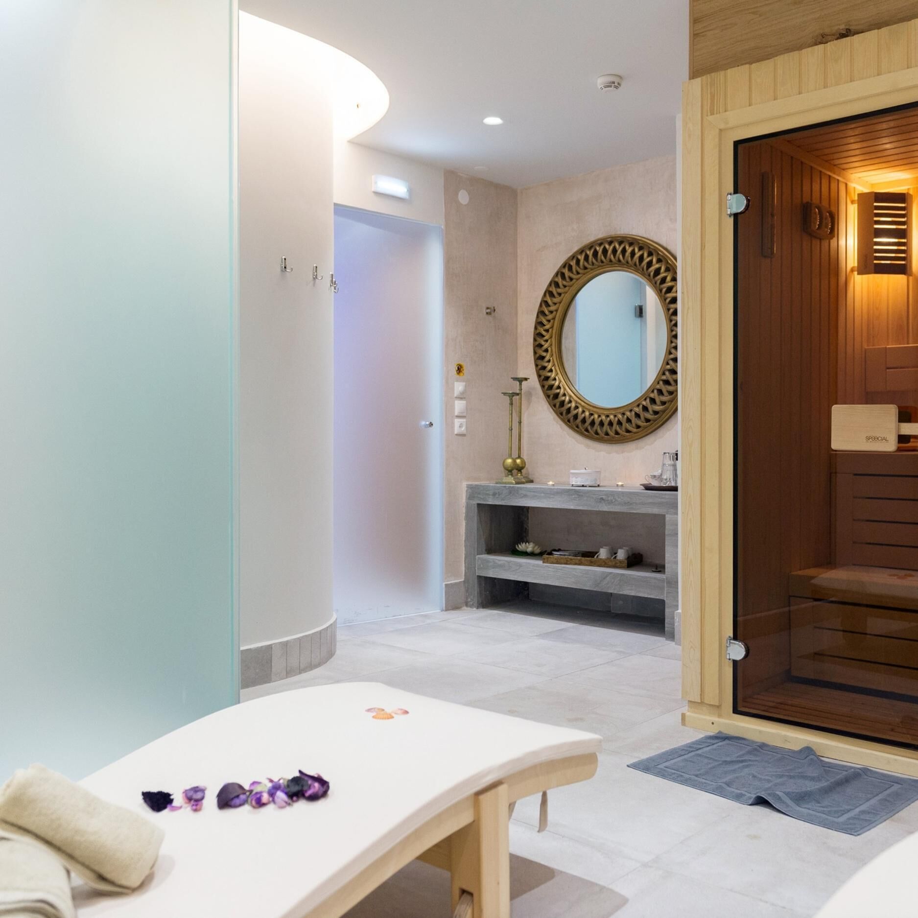 Une salle de spa équipée d’une table de massage, d’un miroir et d’une porte en bois donnant accès à un espace sauna.