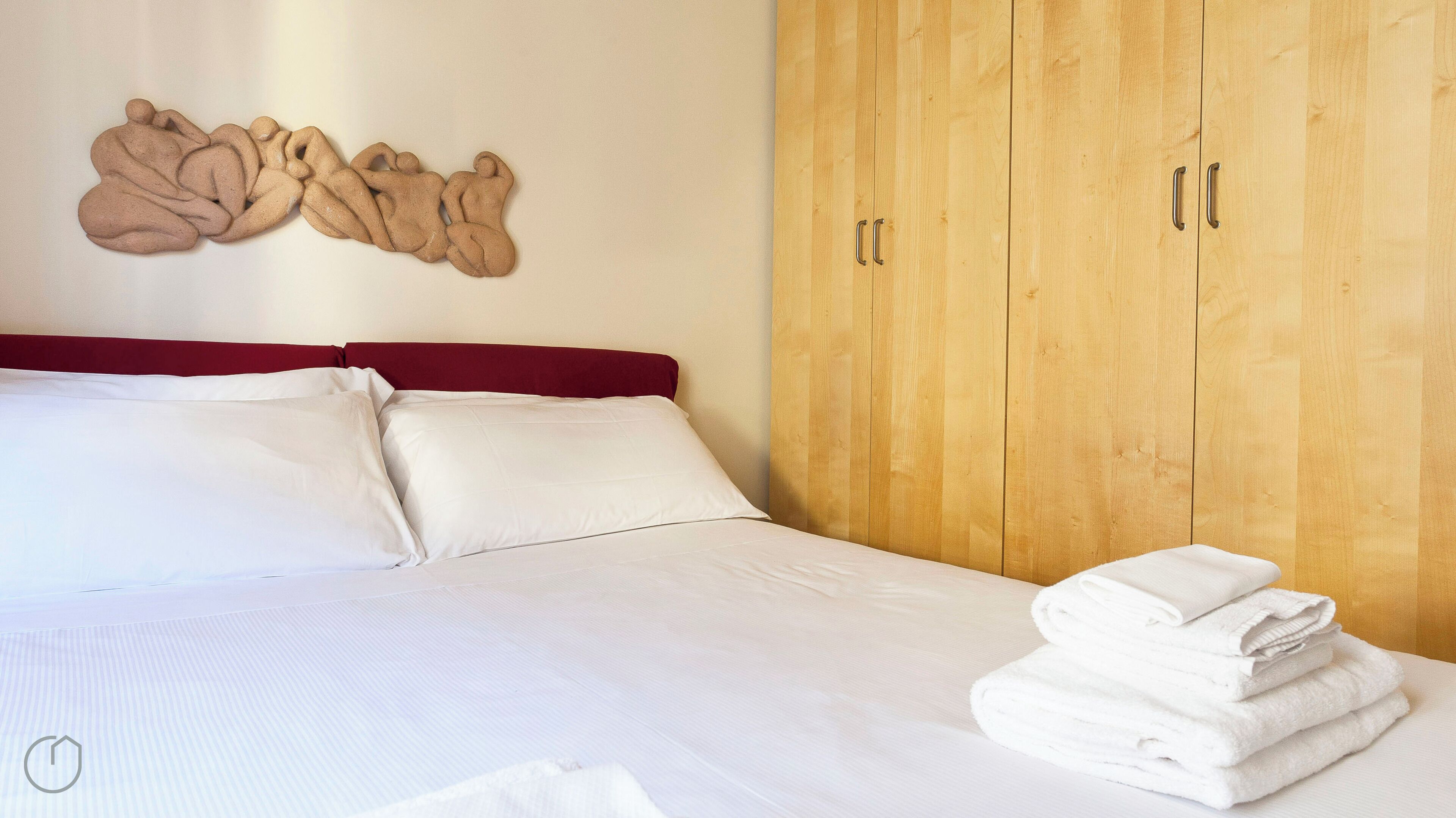A neatly made bed with white linens and a red headboard, a wooden wardrobe with two doors, and a wall decoration.