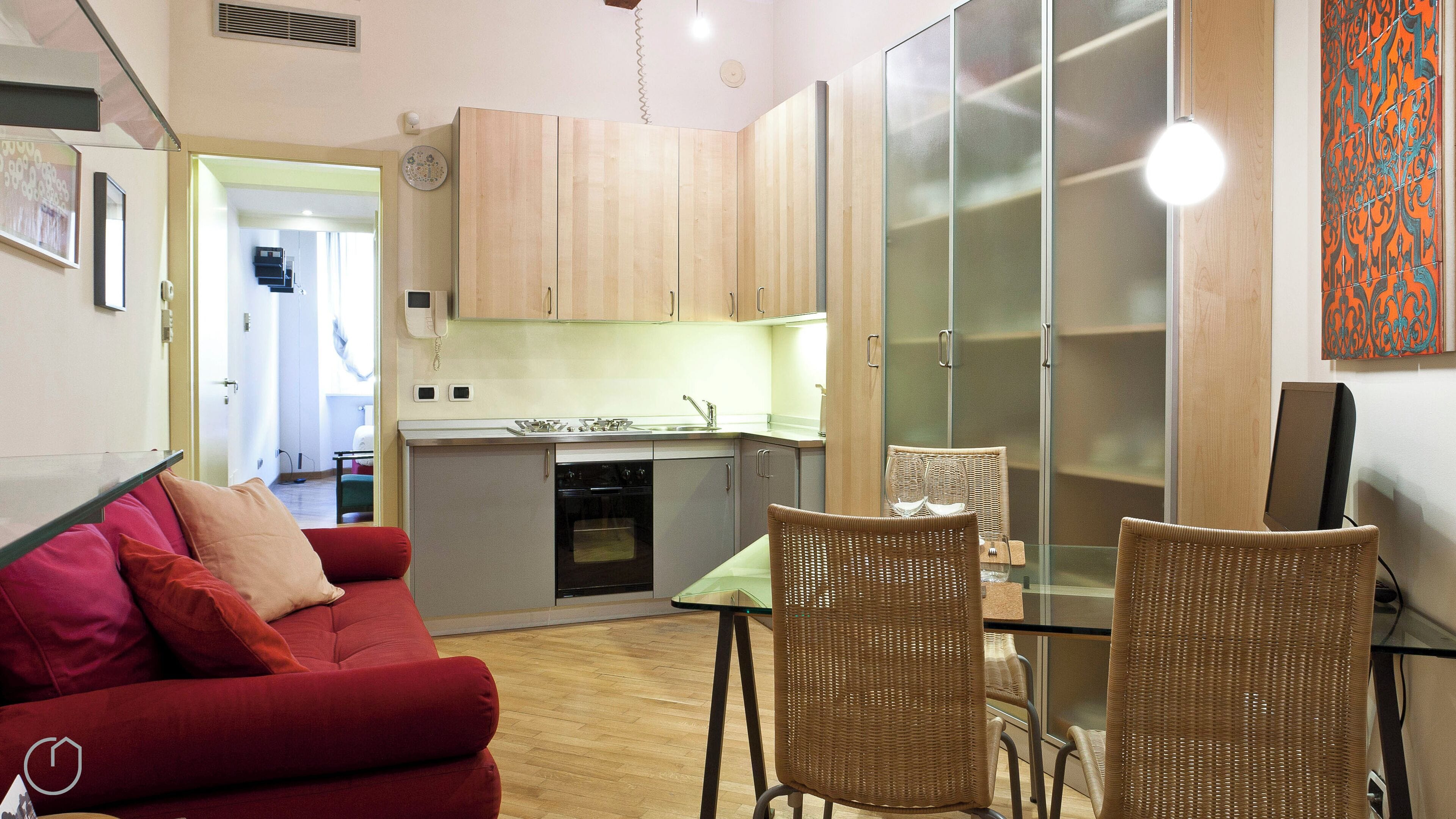Un espace de vie compact comprenant un coin cuisine, un coin repas avec des chaises en osier et un canapé rouge.