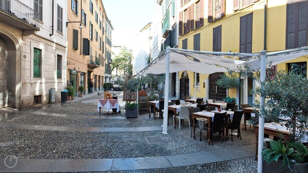 An outdoor café with tables and chairs, surrounded by buildings with balconies and shutters. - Italianway - Fiori Chiari (Milan)