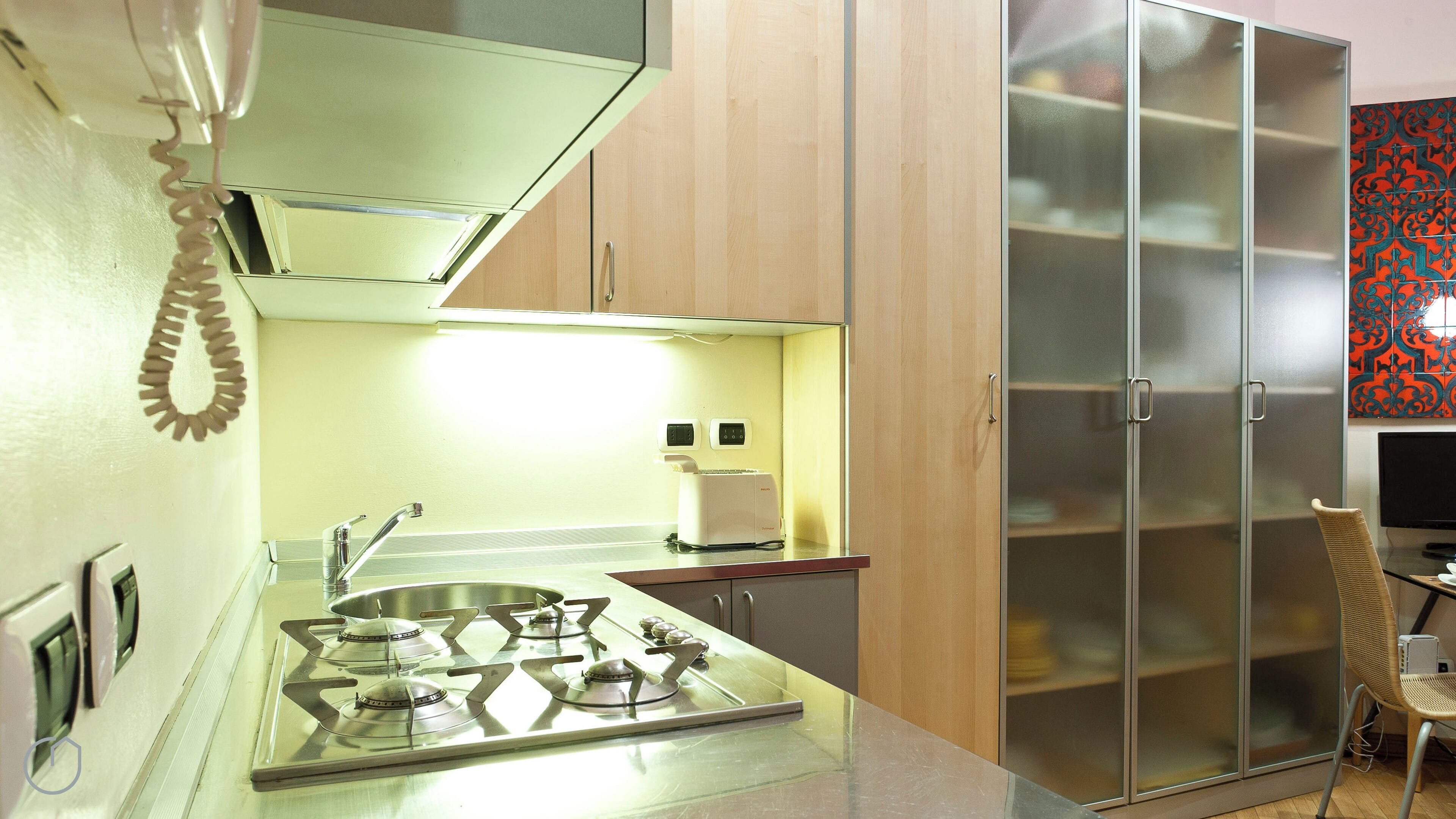 Cozinha compacta com fogão a gás, torradeira e micro-ondas. Há um armário com porta de vidro e uma cadeira no canto.