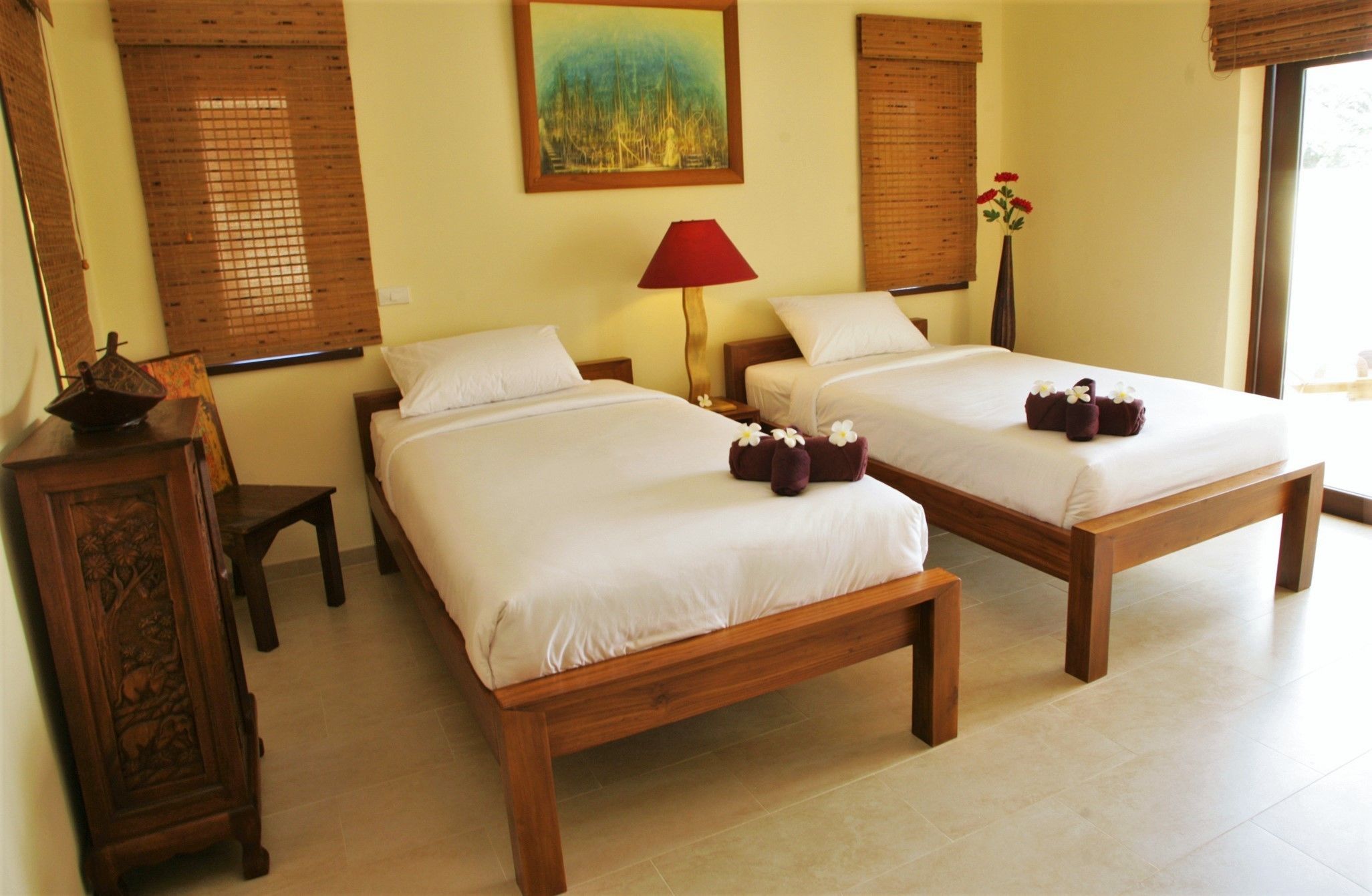 A hotel room with two wooden beds, a red lamp, a painting on the wall, and a window with a view.