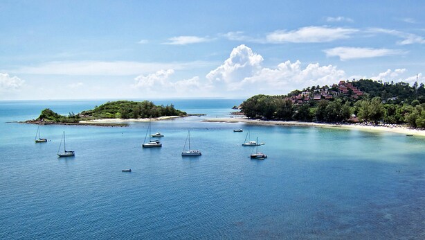 A coastal area with multiple sailboats anchored in calm waters, a sandy beach, and a hillside with buildings. - Chor Chang Villa Resort (Koh Samui)