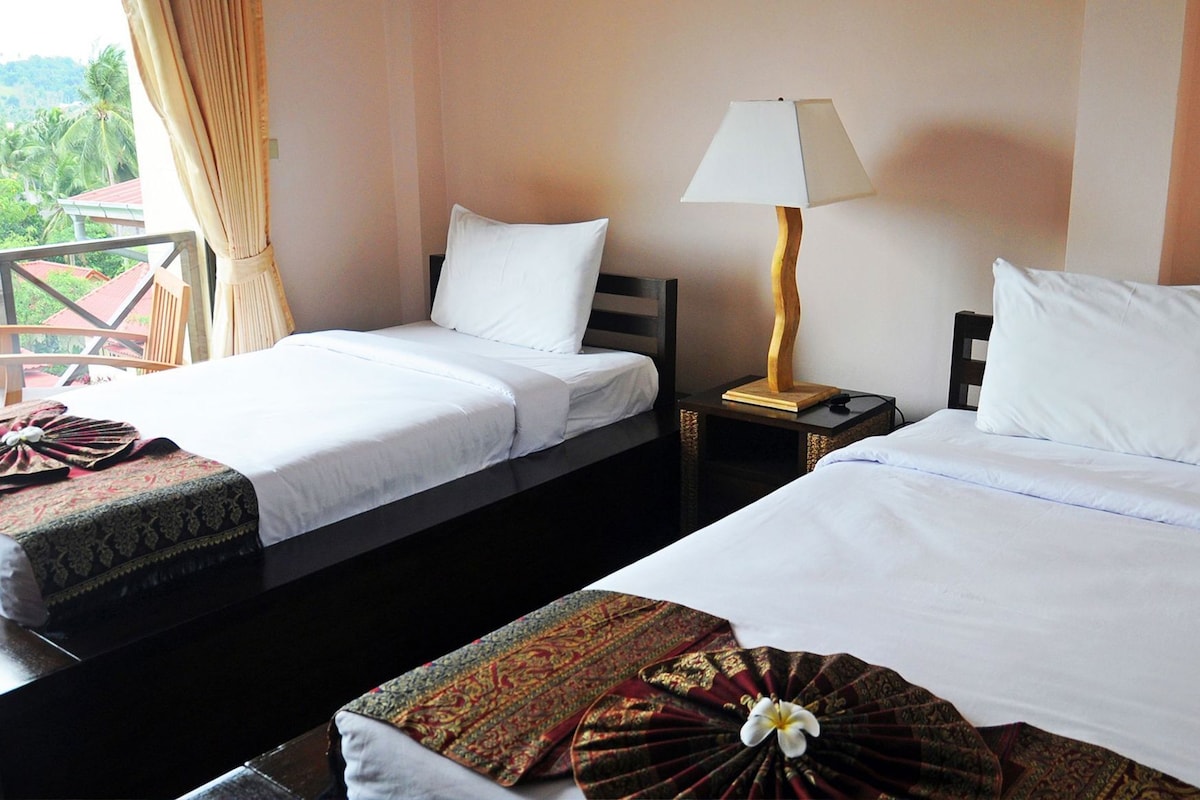 Two single beds with white linens, a wooden nightstand with a lamp, and a balcony with curtains.
