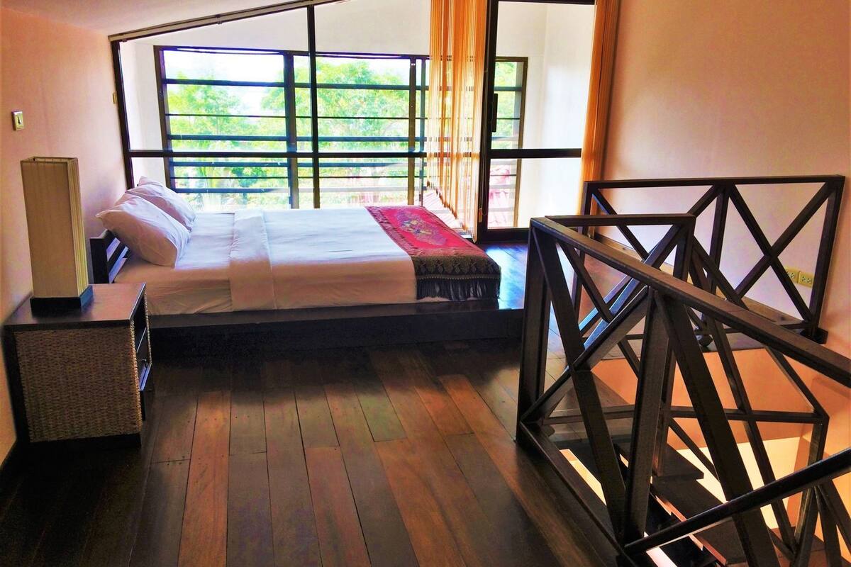A bedroom with a bed, a bedside table, a window with a view of greenery, and a wooden floor.