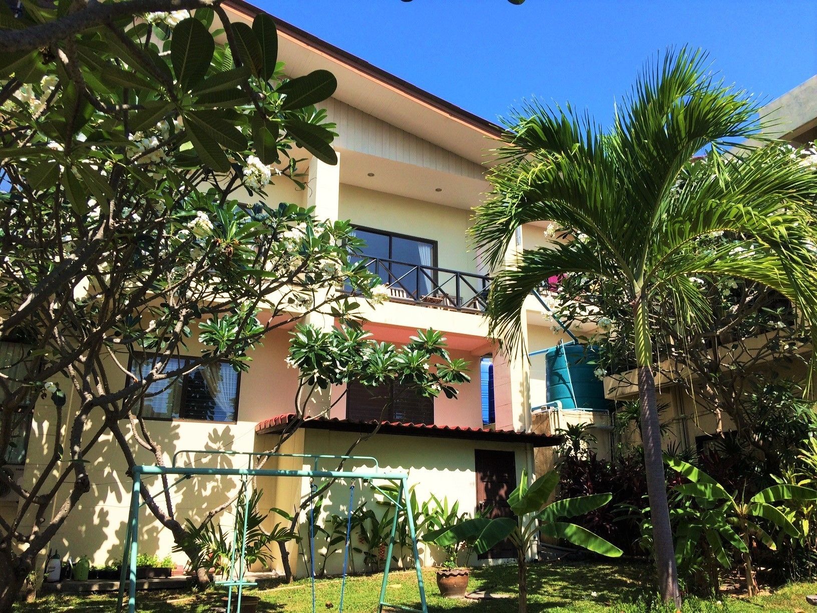 A two-story building with a balcony on the upper floor, surrounded by lush greenery and various plants.