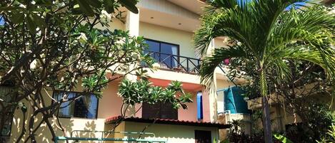 A two-story building with a balcony on the upper floor, surrounded by lush greenery and various plants.