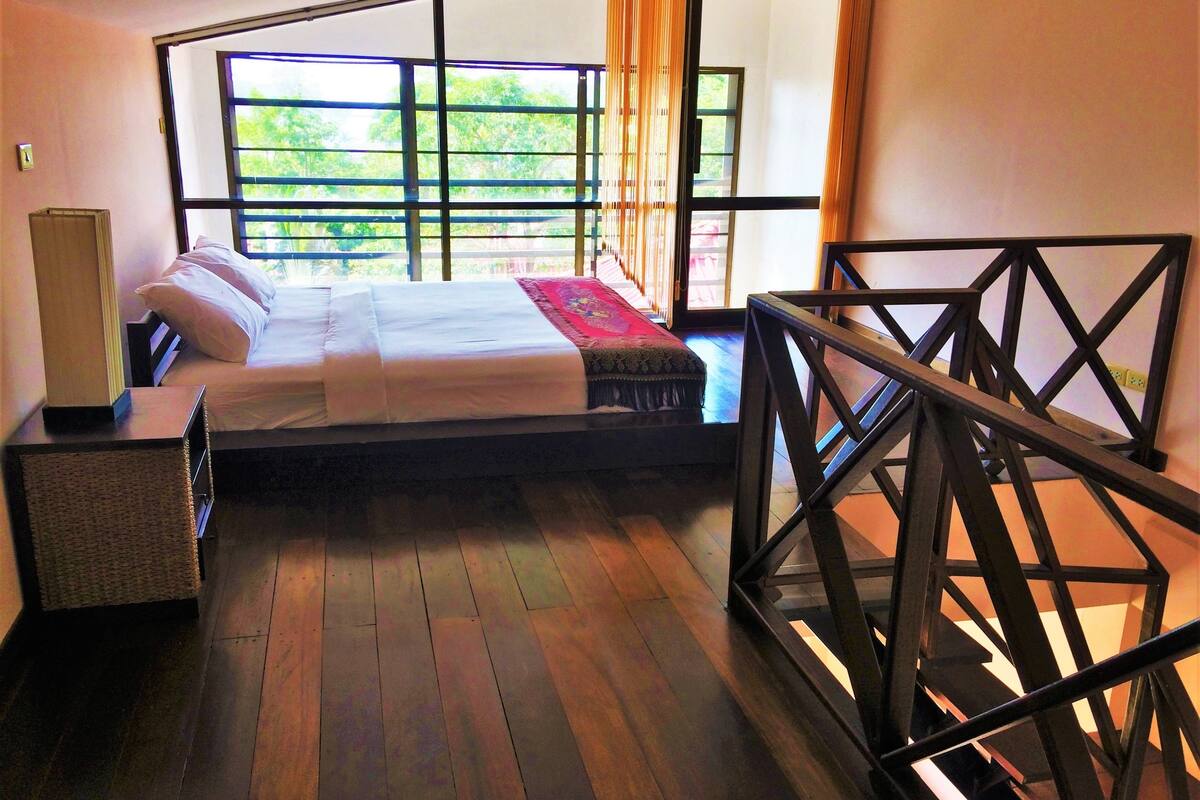 A room with a bed, a bedside table, a lamp, and a view of greenery through large windows.