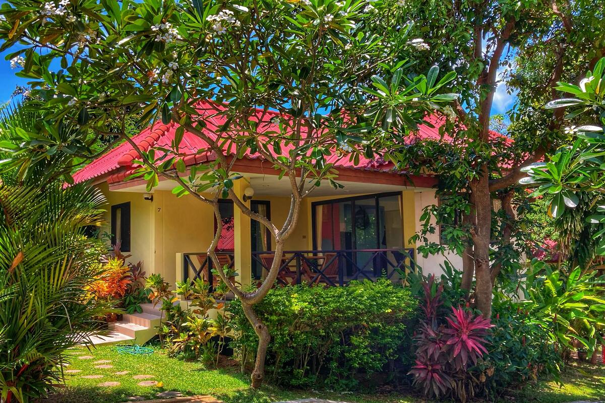 A yellow building with a red roof, surrounded by lush greenery and a small staircase leading to the entrance.