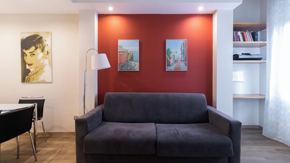 A modern living room with a grey sofa, a dining table with chairs, and two paintings on the wall.