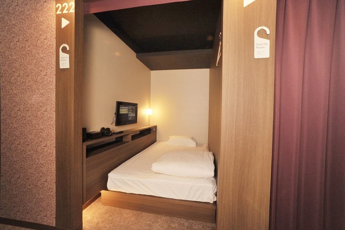 A compact hotel room with a bed, a bedside table, a small shelf, and a wall-mounted TV.