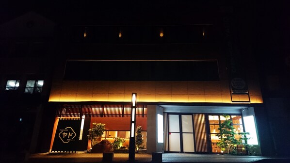 Un edificio con una entrada cálidamente iluminada, un letrero con caracteres japoneses y un pequeño árbol en la parte delantera.