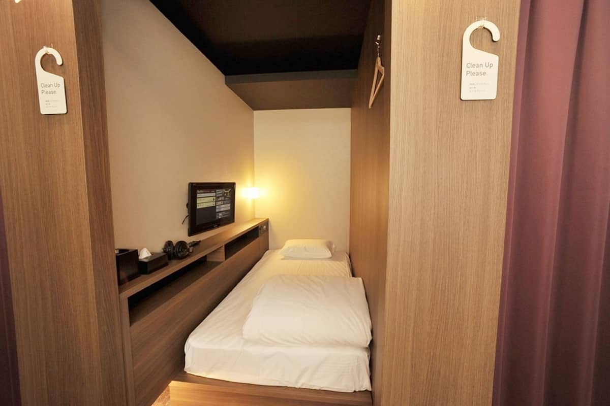 A small, compact room with a single bed, a bedside table with a radio, and a 'Clean Up Please' sign on the door.