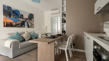 Un appartement moderne comprenant un coin repas, un canapé avec des coussins, une cuisine avec des meubles blancs et un grand tableau représentant un paysage urbain accroché au mur.
