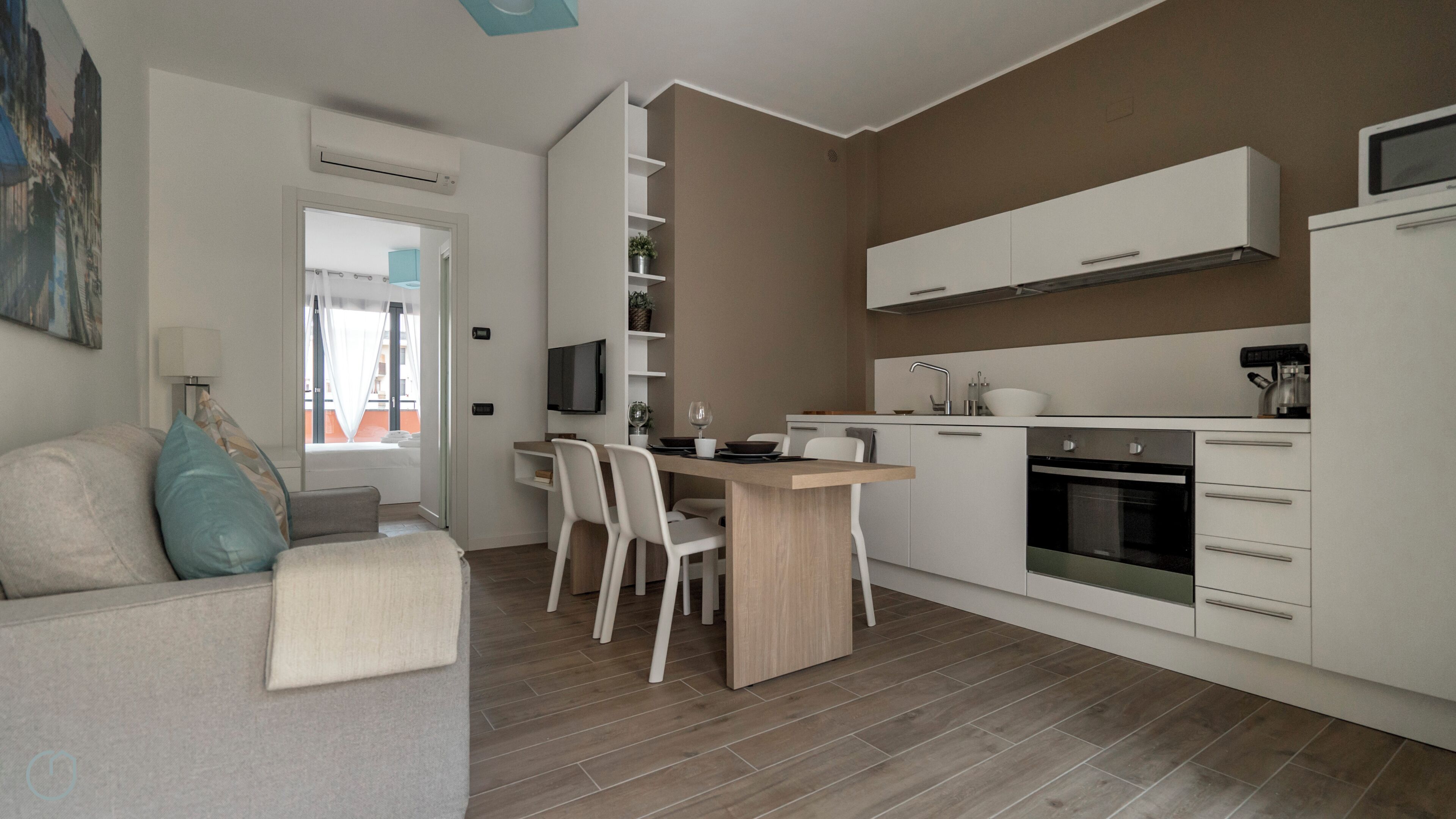 A modern kitchen with white cabinetry, a dining area with a wooden table, and a grey sofa with a blue pillow.