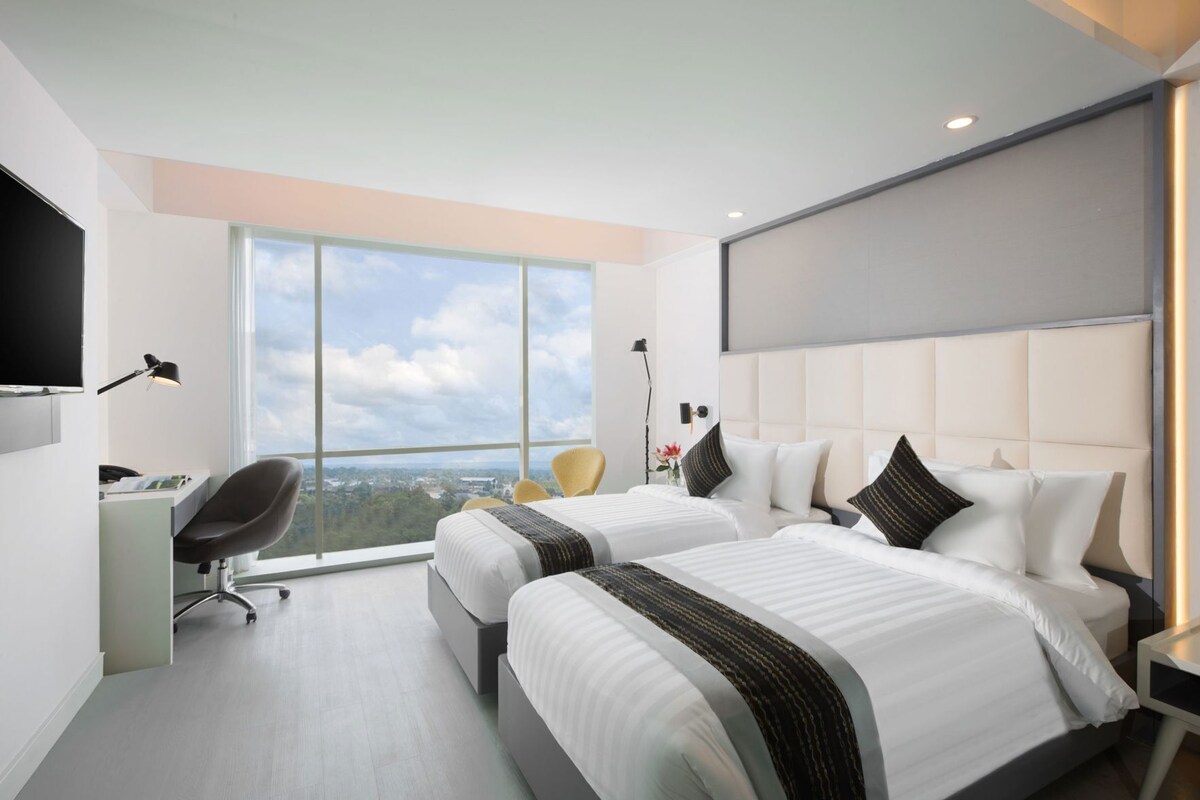 Ett modernt hotellrum med två sängar, ett skrivbord, en stol och ett stort fönster med utsikt över staden.