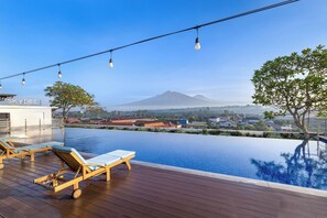 Piscina infinita con terraza de madera y sillones de descanso, con vistas a una montaña y un pueblo.