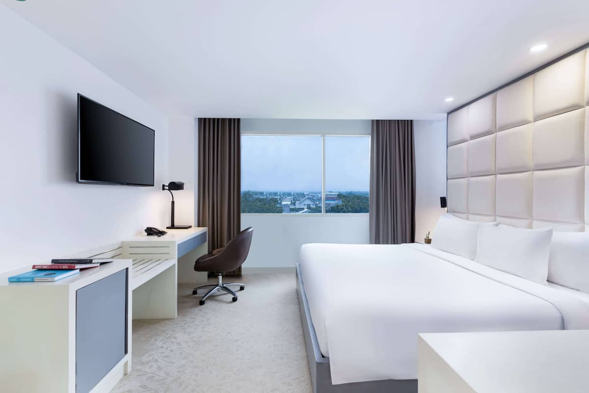 Une chambre d’hôtel moderne dotée d’un grand lit, d’un bureau avec un ordinateur, d’une chaise, d’un téléviseur fixé au mur et d’une fenêtre donnant sur la ville.