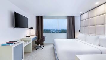 Una habitación de hotel moderna con una cama grande, un escritorio con computadora, una silla, un televisor montado en la pared y un ventanal con vista a la ciudad.