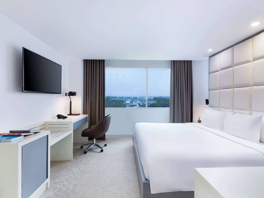 A modern hotel room with a large bed, a desk with a computer, a chair, a TV mounted on the wall, and a window with a city view.