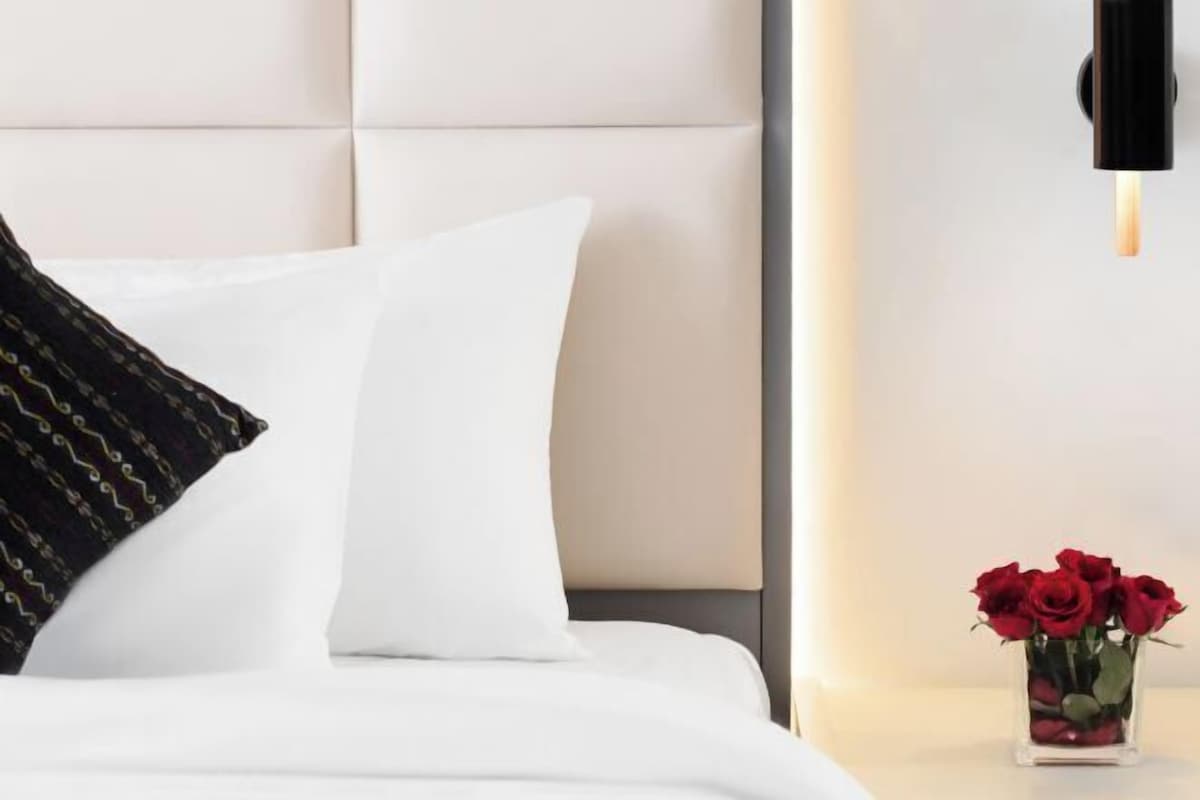 Un lit avec une literie blanche, un coussin noir à motifs et un vase contenant des roses rouges sur une table de chevet.