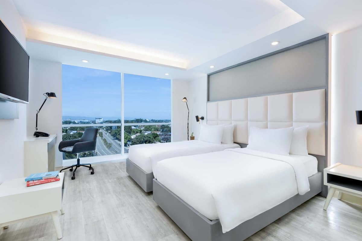 Une chambre d’hôtel moderne avec deux lits, un bureau et une grande fenêtre donnant sur la ville.