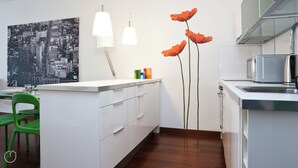 Eine moderne Küche mit weißen Schränken, einem grünen Stuhl, einem Stadtbildgemälde und einer hohen Vase mit orangefarbenen Blumen.